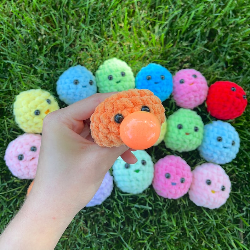May include: A collection of colourful crocheted plush toys with various facial expressions. The toys are arranged in a circle on a green grassy surface. One toy is orange with a large orange nose.