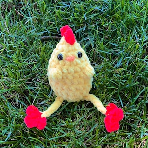 May include: A yellow crocheted chick toy with red feet and a red comb. The chick is sitting on a bed of green grass.
