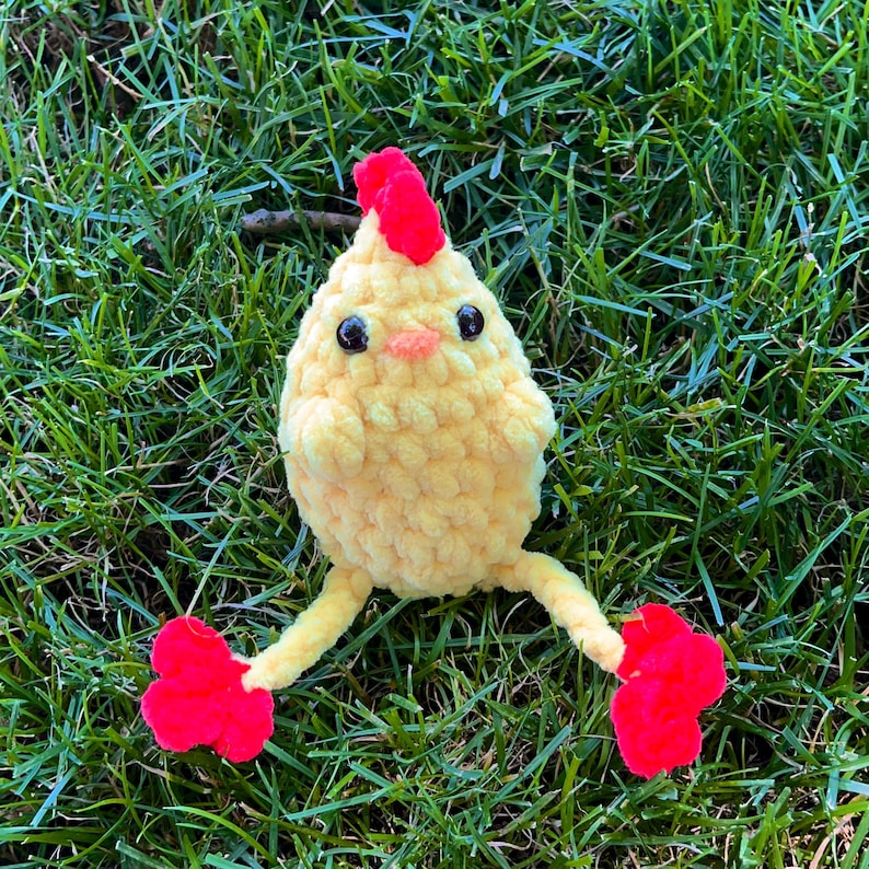 May include: A yellow crocheted chick toy with red feet and a red comb. The chick is sitting on a bed of green grass.
