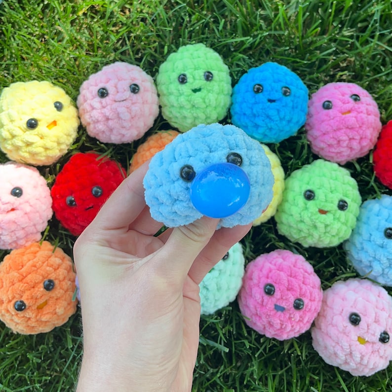 May include: A collection of colourful crocheted plush toys with various facial expressions. The toys are made of soft yarn and have a variety of colours, including pink, blue, green, yellow, and red. One of the toys has a blue dummy.