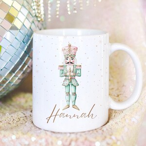 Puede incluir: Taza de cerámica blanca con una ilustración de cascanueces y el nombre "Hannah" escrito en cursiva marrón. El cascanueces lleva un traje rosa y verde y sostiene un bastón dorado.