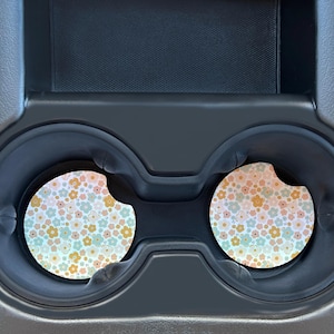 May include: Two round car coasters with a floral pattern in shades of pink, yellow, and blue. The coasters are in a car cup holder.