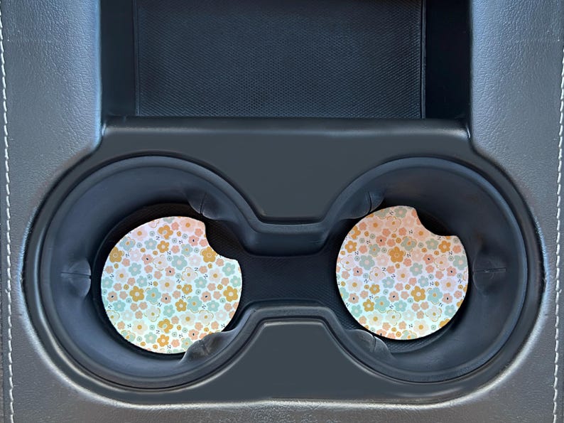 May include: Two round car coasters with a floral pattern in shades of pink, yellow, and blue. The coasters are in a car cup holder.