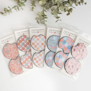 May include: Six sets of car coasters featuring a variety of designs. Each set contains two coasters. The designs include a chequered pattern, a blue and white pattern, a pink and white pattern, and a pattern with smiley faces. The coasters are labelled 'Car Coasters' and 'Handmade with love'.