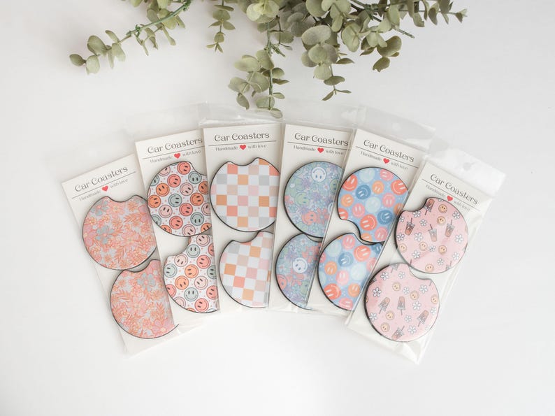 May include: Six sets of car coasters featuring a variety of designs. Each set contains two coasters. The designs include a chequered pattern, a blue and white pattern, a pink and white pattern, and a pattern with smiley faces. The coasters are labelled 'Car Coasters' and 'Handmade with love'.
