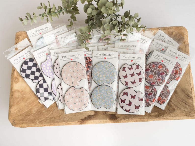 May include: An assortment of car coasters featuring a variety of designs, including floral patterns, butterflies, and a checkered pattern. The coasters are packaged in clear plastic bags with the text 'Car Coasters' printed on them.