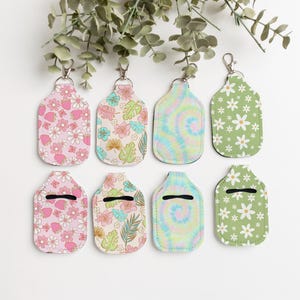 May include: Eight hand sanitiser holders with different patterns. The patterns include pink flowers, green leaves, and tie-dye. The holders are all attached to silver keychains.