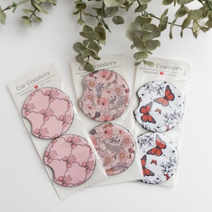 May include: Set of six car coasters featuring a variety of floral and butterfly designs. The coasters are pink, white and red with black outlines. The packaging reads "Car Coasters Handmade with Love".