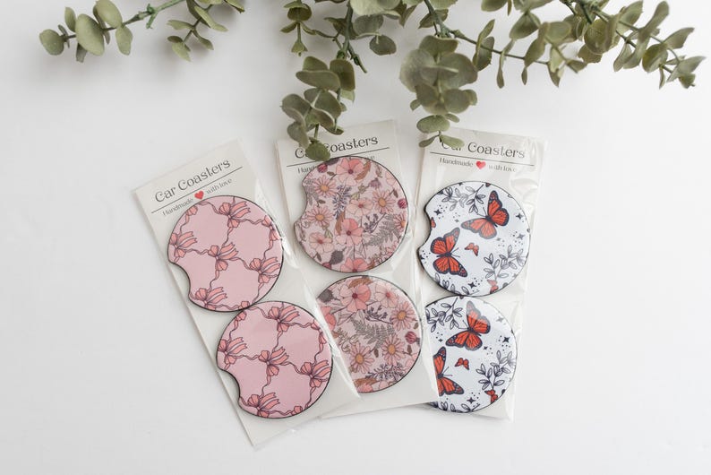 May include: Set of six car coasters featuring a variety of floral and butterfly designs. The coasters are pink, white and red with black outlines. The packaging reads "Car Coasters Handmade with Love".