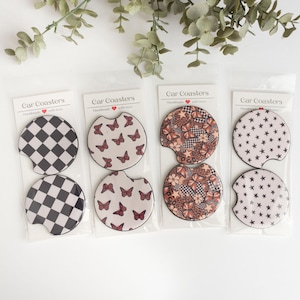 May include: Set of four car coasters featuring different designs. The coasters are round and have a white background with various patterns in black, brown, and red. The patterns include butterflies, stars, and chequered designs. The coasters are packaged in clear plastic sleeves with the text "Car Coasters" printed on the front.