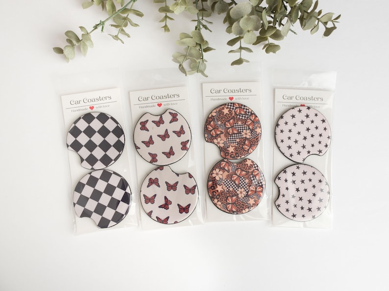 May include: Set of four car coasters featuring different designs. The coasters are round and have a white background with various patterns in black, brown, and red. The patterns include butterflies, stars, and chequered designs. The coasters are packaged in clear plastic sleeves with the text "Car Coasters" printed on the front.