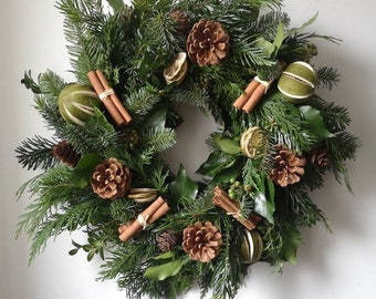 May include: A festive wreath composed of evergreen branches, pinecones, dried citrus slices, and cinnamon sticks. The wreath is circular and features a variety of green foliage, with brown pinecones and cinnamon sticks adding warmth. The dried citrus slices are a light green color.