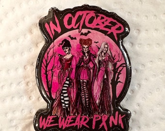 May include: A black and pink pin with the text "IN OCTOBER WE WEAR PINK". The pin features a pink background with three illustrated figures in dresses, bats, and bare trees.