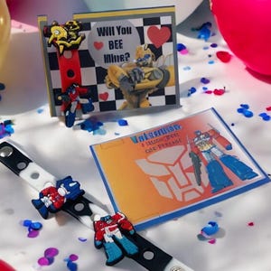 Könnte beinhalten: Eine Valentinstags-Ausstellung mit Accessoires im Transformers-Stil. Enthält ein Armband mit Charakter-Anhängern, Grußkarten mit dem Text "Will You BEE Mine?" und "Transformers" sowie verstreute Konfetti.