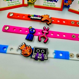 May include: Three colorful silicone bracelets with different cartoon character charms. The bracelets are orange, pink, and blue and white. The charms include a monster, a frog, a robot, and a character from the show "Rainbow Friends".