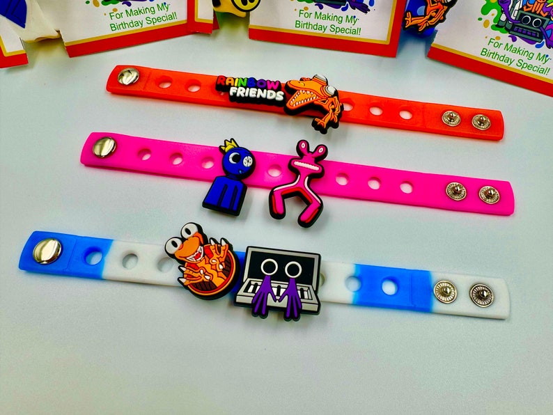 May include: Three colorful silicone bracelets with different cartoon character charms. The bracelets are orange, pink, and blue and white. The charms include a monster, a frog, a robot, and a character from the show "Rainbow Friends".