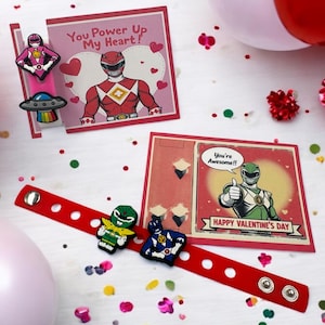 Könnte beinhalten: Artikel zum Valentinstag. Ein rotes Armband mit Power Ranger-Anhängern, Karten mit Power Rangers-Illustrationen und den Texten "You Power Up My Heart!" und "Happy Valentine's Day". Konfetti und Luftballons.