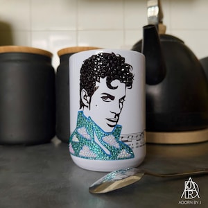May include: White ceramic mug featuring a black and white portrait with a teal and white cloud design. The hair is embellished with black crystals. A silver spoon with matching embellishments is in front of the mug.