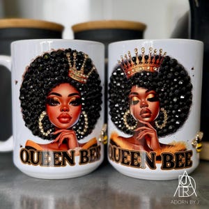 May include: Two white ceramic mugs with a "Queen Bee" design. Each mug features a portrait of a woman with a jeweled afro, crown, and gold hoop earrings. The text "Queen Bee" is printed on the front.