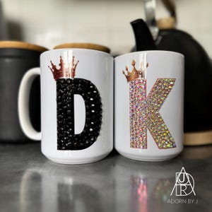 May include: Two white ceramic mugs with the letters "D" and "K" embellished with black and iridescent rhinestones. Each mug features a gold crown design at the top. The mugs are part of a set.
