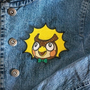 May include: A blue denim jacket with a yellow, brown and green embroidered patch of an owl with a bow tie. The owl is surrounded by a yellow starburst.