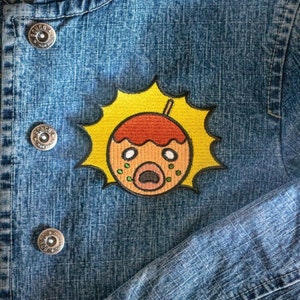 May include: A yellow sunburst with a cartoon face inside. The face is orange with red hair and green spots. The face is making a surprised expression with its mouth open.