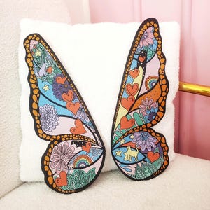 Peut inclure: Deux ailes de papillon colorées avec un design fantaisiste comprenant des cœurs, des étoiles, des arcs-en-ciel et des fleurs. Les ailes sont peintes dans des tons de rose, bleu, jaune, vert et orange.
