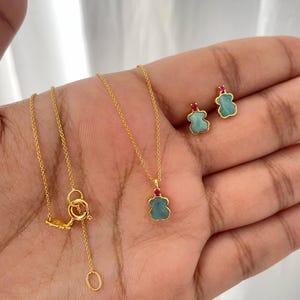 May include: A gold-toned necklace and matching earrings set. The necklace features a small pendant with a turquoise-colored bear-shaped stone and a red accent. The earrings also have turquoise bear-shaped stones with red accents.