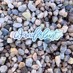 May include: A rainbow-colored vinyl sticker with the word "Wanderlust" written in a cursive font. The sticker is placed on a bed of small, gray, white, and brown pebbles.
