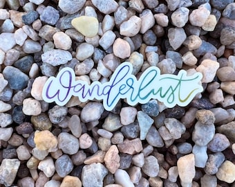 May include: A rainbow-colored vinyl sticker with the word "Wanderlust" written in a cursive font. The sticker is placed on a bed of small, gray, white, and brown pebbles.