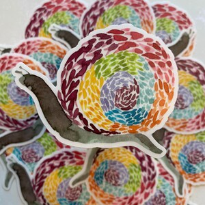 May include: A stack of colorful snail stickers with a rainbow shell design. The snail is painted in a watercolor style with a brown body and a shell with a swirling pattern of red, orange, yellow, green, blue, purple, and pink.