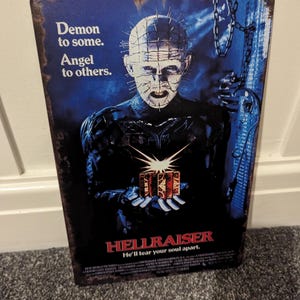 May include: A vintage style metal sign featuring a character from the horror movie Hellraiser. The character is a demon with a hooked chain and a box in his hand. The text on the sign reads "Demon to some. Angel to others. Hellraiser. He'll tear your soul apart."