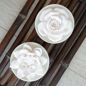 May include: Two white ceramic diffuser flower tops. The flower tops are in the shape of a rose and a lotus flower. The flower tops are sitting on a brown bamboo mat.