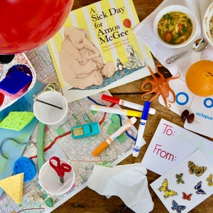 Puede incluir: Una mesa con una variedad de artículos, incluyendo un libro titulado "A Sick Day for Amos McGee", un globo rojo, un plato de avena, un tazón de sopa, un pulpo de juguete, un coche de juguete, marcadores, esponjas, pintura y una tarjeta con las palabras "To: Miss Ali" y "From:".