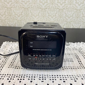 Puede incluir: Radio reloj digital FM/AM Sony Dream Machine ICF-C122 negro. El radio reloj tiene una pantalla digital y botones en la parte superior. La radio tiene bandas de radio AM y FM.