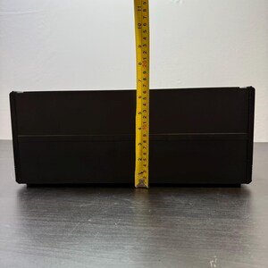 May include: A black rectangular box with a yellow measuring tape next to it. The tape shows the box is approximately 10 inches tall. The box is on a dark wooden surface.