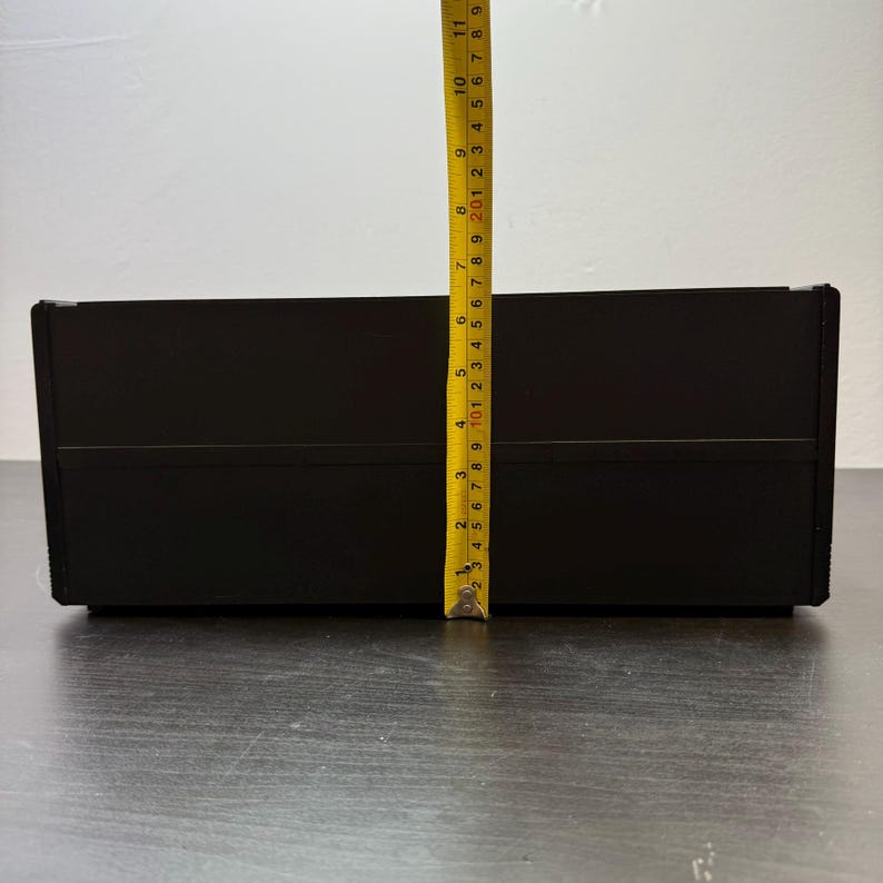 May include: A black rectangular box with a yellow measuring tape next to it. The tape shows the box is approximately 10 inches tall. The box is on a dark wooden surface.