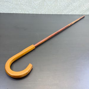 Puede incluir: Un bastón largo y delgado que presenta un mango de madera clara en forma de J, unido a un eje de madera de color marrón rojizo más oscuro con un patrón de veta natural. El bastón es recto y se estrecha ligeramente hacia el extremo.