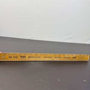 May include: A vintage wooden ruler with inch markings and text. The ruler is light brown with black text that reads "HA. 3251" and "Impressions - Gravure - Reliure - Reglage". The ruler is approximately 30 cm long.