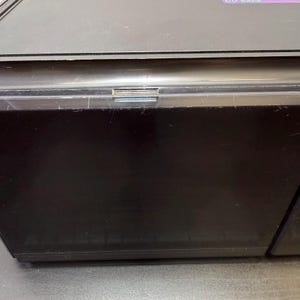 May include: A black electronic device with a hinged lid. The device has a rectangular shape and appears to be made of plastic. The lid has a thin, silver-colored strip along the top edge. The device is sitting on a dark surface.