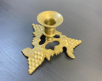 May include: A gold metal candle holder with a grapevine design. The holder has a round top and a base with three grapevine branches.