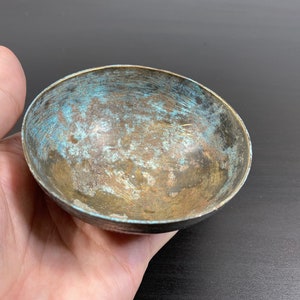 May include: A small, round, metal bowl with a worn, aged patina. The bowl has a light blue and brown colour with a textured surface.