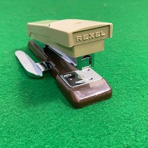May include: A brown and beige Rexel stapler with a silver lever and a built-in staple remover. The stapler is on a green surface.