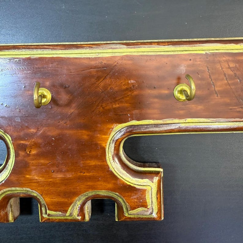 May include: Wooden coat rack for wall mounting, featuring two brass hooks. The rack has a dark brown finish with gold trim and decorative cutouts. A vintage-style piece for a hallway or bedroom.