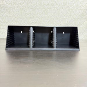 May include: A black plastic media storage rack featuring three distinct compartments separated by vertical dividers with horizontal ridges. The unit has two keyhole slots on the back panel for wall mounting and a rectangular frame design.