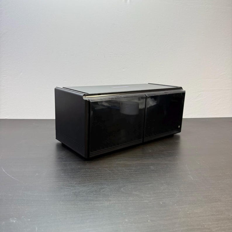 May include: A black, rectangular audio component with a glossy finish. The device has two compartments with a clear, transparent cover. The top of the component is also black and glossy. The device is sitting on a dark surface.