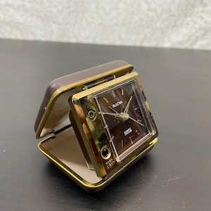 May include: A portable Bulova quartz travel alarm clock with a brown square face and gold-tone metal frame. The clock is housed in a hinged brown case with a light-colored interior. The face features the text Bulova and Quartz Lite Alarm.