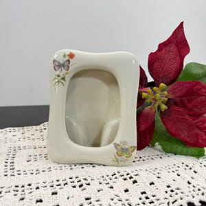 May include: Off-white ceramic picture frame with a curved rectangular shape. The frame is decorated with butterfly and floral designs in pastel colors. A red poinsettia flower is in the background.