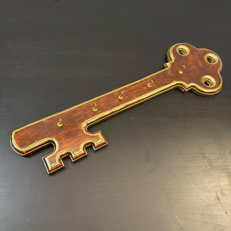 May include: A decorative key-shaped wall-mounted key holder. The key holder is made of dark brown wood with a gold-coloured trim and key hooks. The key holder is approximately 30 cm long.