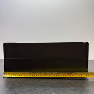 May include: A black rectangular box with a yellow measuring tape laid across the bottom. The tape shows a measurement of approximately 15 inches. The box is on a dark surface with a white background.